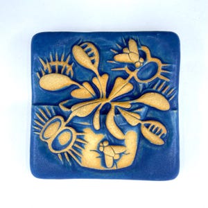 May include: A square, blue ceramic tile with a raised design of a Venus flytrap and insects. The flytrap and insects are tan, contrasting with the blue background. The tile is a decorative piece, possibly for a wall or tabletop.