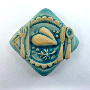 May include: A turquoise ceramic tile with a raised design of a plate setting. The plate features a beige food item, a fork, knife, and spoon. The tile has a diamond shape with a scalloped edge and is suitable for kitchen decor.