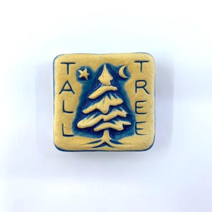May include: A square ceramic tile with rounded corners featuring a debossed evergreen tree design in deep blue against a textured tan background. The text TALL TREE is vertically oriented on both sides of the tree, with a small star and crescent moon.