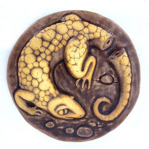 May include: A ceramic wall plaque featuring a brown and yellow lizard with a textured surface. The lizard is curled up in a circle with its tail wrapped around its body. The plaque has a brown background with a few small white dots.