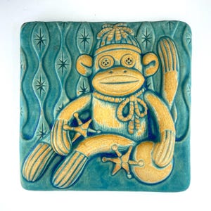 May include: A ceramic tile featuring a sock monkey design. The monkey is in shades of yellow and tan, wearing a hat and scarf, with button eyes. The background is teal with a repeating star and leaf pattern.