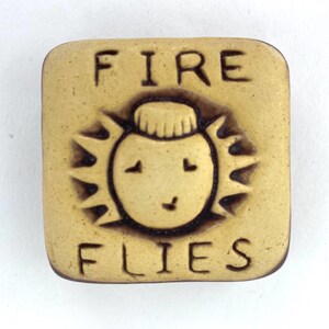 May include: A square, tan-colored ceramic tile with the words "FIRE FLIES" in dark brown lettering. The center features a smiling sun-like face with spiky rays. The design is embossed, giving it a textured appearance.
