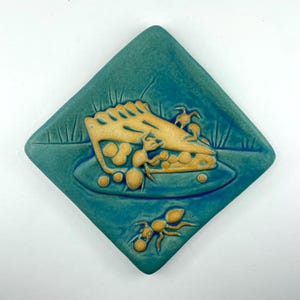 May include: A teal ceramic tile with a raised relief design of a pie slice and ants. The pie is tan with a golden crust, and the ants are also tan. The tile is square with rounded corners and a textured surface.