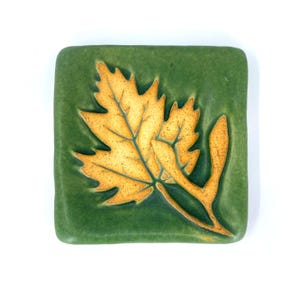 May include: A square ceramic tile with a matte green finish featuring a raised, embossed golden-yellow maple leaf design. The leaf has detailed veins and a textured surface, centered on the tile with rounded corners and smooth edges.