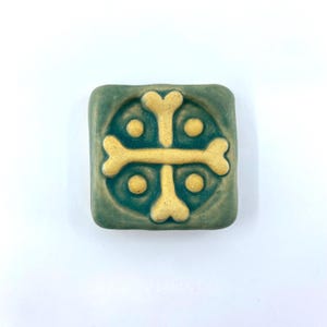 May include: A square ceramic tile with a matte teal green finish featuring a raised, light yellow cross design composed of four bone-shaped segments. Four small, raised yellow dots are positioned in the quadrants between the cross arms.