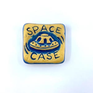 May include: A square ceramic tile with rounded corners featuring a raised gold-colored UFO graphic in the center. The text SPACE is embossed above the UFO and CASE is embossed below it, all set against a deep blue background with gold borders.