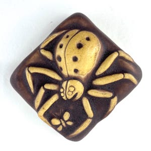 May include: A square, brown decorative button with a raised design of a yellow spider and a small butterfly. The spider has a yellow body with black spots and long, yellow legs. The button is likely made of ceramic or a similar material.
