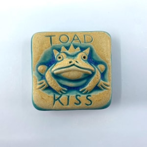 May include: Square ceramic tile with a raised design of a crowned toad. The toad is tan with blue accents, and the tile has the words "TOAD KISS" in a matching blue color.