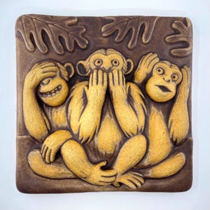 May include: A square, brown ceramic tile featuring a relief sculpture of the three wise monkeys: see no evil, hear no evil, speak no evil. The monkeys are in a seated position with their hands covering their eyes, ears, and mouth. The monkeys are a light tan color.
