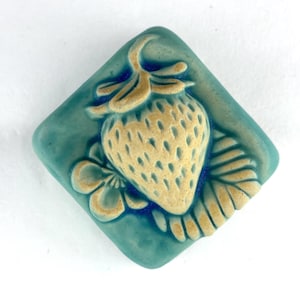 May include: A square, turquoise ceramic tile with a raised strawberry design. The strawberry is beige with a textured surface, and is surrounded by a flower and leaves. The tile has a diamond shape.