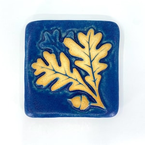 May include: A square ceramic tile with a deep blue glazed finish featuring a raised, embossed design of two light yellow oak leaves and a single acorn. The surface has a smooth, glossy texture with subtle variations in the blue color depth.