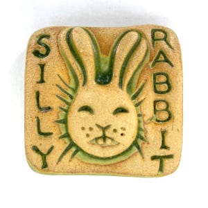 May include: A square ceramic tile with a rabbit design. The tile is tan with a green rabbit and the words "SILLY RABBIT" in green. The rabbit has a smiling face and long ears. The tile is likely handmade.