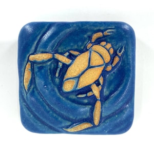 May include: A blue ceramic tile with a raised design of a water beetle in a swirling water pattern.
