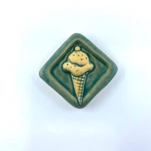 May include: A diamond-shaped ceramic tile featuring a raised, textured yellow ice cream cone design centered on a matte, dark green background. The piece has a smooth, glazed finish with defined edges and a relief pattern of a scoop on a cone.