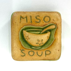 May include: Square ceramic tile with a miso soup design. The tile features the words "MISO SOUP" in a dark green color. The soup bowl is light yellow with a green border.