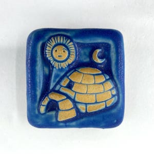 May include: Square blue ceramic tile featuring a stylized igloo, sun, and crescent moon design. The igloo is tan, and the sun has a smiling face. The background is a darker blue with a lighter blue border.