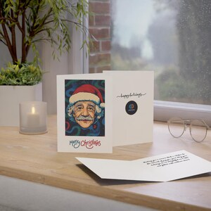 May include: A Christmas card featuring a colorful illustration of Albert Einstein wearing a Santa hat. The card reads "Merry Christmas" and includes a small logo for the company "Adventure" in the bottom right corner.
