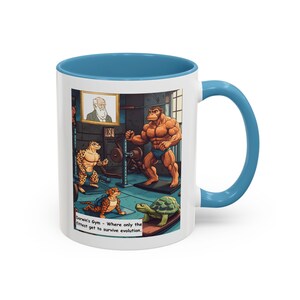 Puede incluir: Taza de café de cerámica blanca con borde y asa de color azul claro. La taza presenta una ilustración de dibujos animados de un simio musculoso levantando pesas en un gimnasio. Otros animales de dibujos animados están presentes, incluyendo una tortuga, un lagarto y un hombre con barba. El texto en la taza dice "Darwin's Gym - Where only the fittest get to survive evolution."