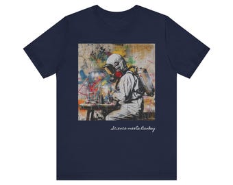 Op de afbeelding: Een marineblauw t-shirt met een grafische print van een persoon die een gasmasker en een wit beschermend pak draagt. De persoon zit aan een tafel met een schaakbord en stukken. De tekst "Science meets Banksy" is onder de afbeelding gedrukt.