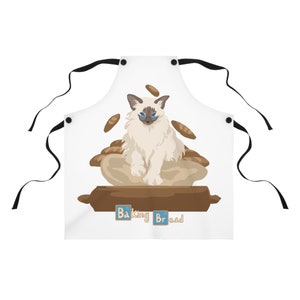 May include: White apron with a white cat wearing blue glasses sitting on a rolling pin with bread around it. The text "Baking Bread" is below the rolling pin.