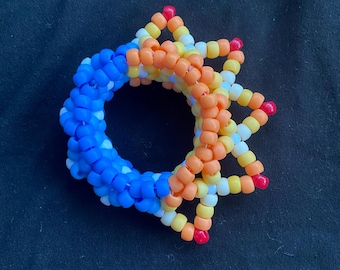 Può includere: Un bracciale circolare realizzato con perline di plastica. Il design presenta un vivace gradiente di colori che passa dal blu intenso all'arancione, al giallo e al bianco, con piccole perline rosse che accentuano le punte esterne.