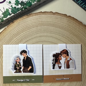Puede incluir: Dos marcapáginas ilustrados con parejas de una novela de fantasía. El primer marcapáginas muestra a una mujer con el pelo largo y blanco y un hombre con el pelo oscuro. El texto debajo dice "Paedyn & Kai". El segundo marcapáginas muestra a una mujer con el pelo largo y castaño y un hombre con el pelo oscuro. El texto debajo dice "Adena & Makoto".