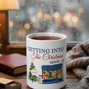 Puede incluir: Una taza de cerámica blanca con el texto 'GETTING INTO The Christmas spirit...' en azul y rojo. El diseño incluye un árbol de Navidad decorado, una librería azul y un pequeño personaje fantasma sosteniendo un libro.