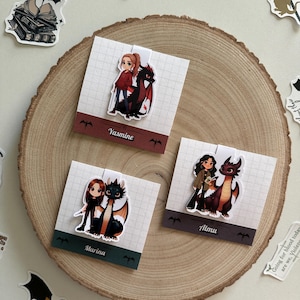 May include: Three colourful bookmarks featuring illustrations of women with dragons. The bookmarks are on a wooden surface. The bookmarks are labelled "Marina", "Yasmine", and "Almu".