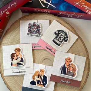 May include: Five different bookmark designs featuring characters from the book "Boys of Tommen." The bookmarks are printed on white cardstock with a grid pattern. The designs include a crest with the words "Tommen College," a jersey with the number "13" and the name "Johnny," and three couples with the names "Shannon & Johnny," "Gibsie & Claire," and "Joey & Aoife."
