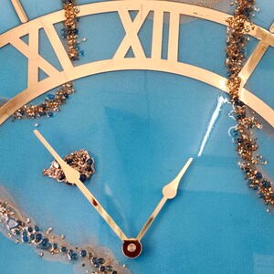 May include: A blue resin clock face with gold Roman numeral hour markers and gold hands. The clock face is decorated with gold glitter and blue and gold beads.