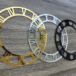 Może przedstawiać: Trzy dekoracyjne kontury tarczy zegara w kolorze złotym, srebrnym i czarnym. Kontury są wykonane z metalu i mają oznaczenia cyfr rzymskimi.