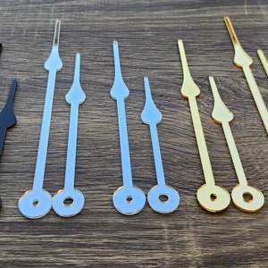 May include: A set of clock hands in black, silver, and gold. The hands are made of metal and have a pointed tip. There is also a clock movement mechanism included.