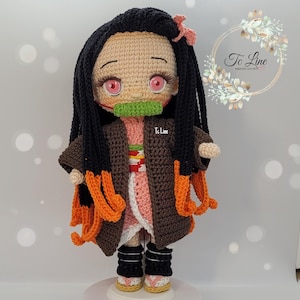 May include: Crocheted doll with black hair, pink cheeks, and a green mouth covering. The doll is wearing a brown and pink kimono with orange accents. The doll is standing on a white surface.