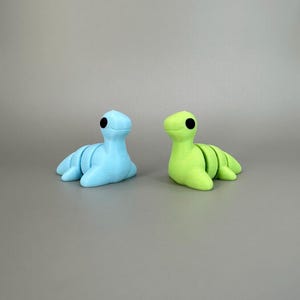 May include: Two small, stylized sculptures of a sea creature, one light blue and the other lime green. Each has a segmented body, flipper-like appendages, and a black dot for an eye. The sculptures are set against a neutral gray background.