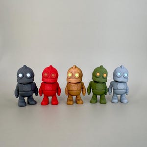 May include: Five small robot figurines in a row, each with a different color: dark gray, red, tan, green, and light gray. Each robot has a round head, two eyes, and a rectangular body with arms and legs. The eyes glow.