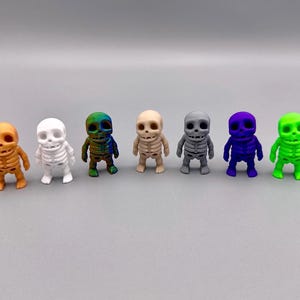 May include: Seven small skeleton figurines in different colors, including orange, white, green, blue, gray, and a multi-colored skeleton. The skeletons are all in the same pose, with their arms outstretched and their legs slightly apart.