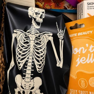 May include: A black pouch featuring a white skeleton graphic making a hand gesture, positioned next to an orange package labeled Taste Beauty Don't Jelly Jelly Sheet Mask Pineapple Scented with a net weight of 2.1 oz, and a lined paper card.