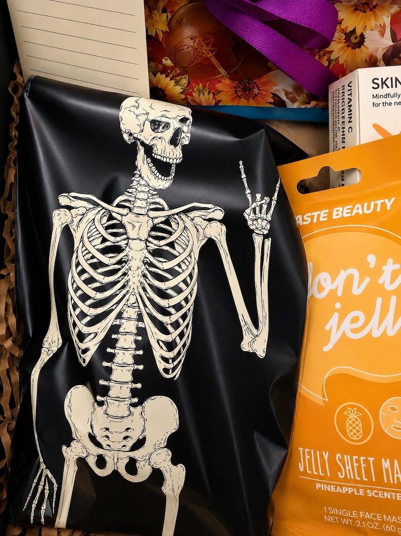 May include: A black pouch featuring a white skeleton graphic making a hand gesture, positioned next to an orange package labeled Taste Beauty Don't Jelly Jelly Sheet Mask Pineapple Scented with a net weight of 2.1 oz, and a lined paper card.