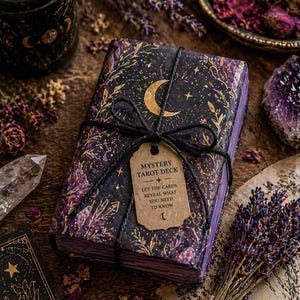 May include: A wrapped tarot deck with a celestial design featuring a crescent moon and stars, tied with black twine. The tag reads "Mystery Tarot Deck" and "Let the cards reveal what you need to know." The deck is surrounded by dried flowers and crystals.