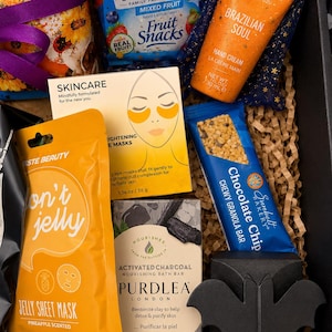 May include: A gift box containing a Welch's mixed fruit snack pack, a Spongelle Brazilian Soul hand cream, a box of brightening eye masks, a Taste Beauty pineapple jelly sheet mask, a Sunbelt Bakery chocolate chip granola bar, and a Purdlea activated charcoal bath bar with a black bat-shaped decorative item.