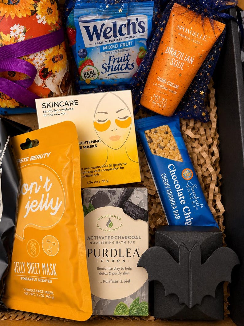 May include: A gift box containing a Welch's mixed fruit snack pack, a Spongelle Brazilian Soul hand cream, a box of brightening eye masks, a Taste Beauty pineapple jelly sheet mask, a Sunbelt Bakery chocolate chip granola bar, and a Purdlea activated charcoal bath bar with a black bat-shaped decorative item.