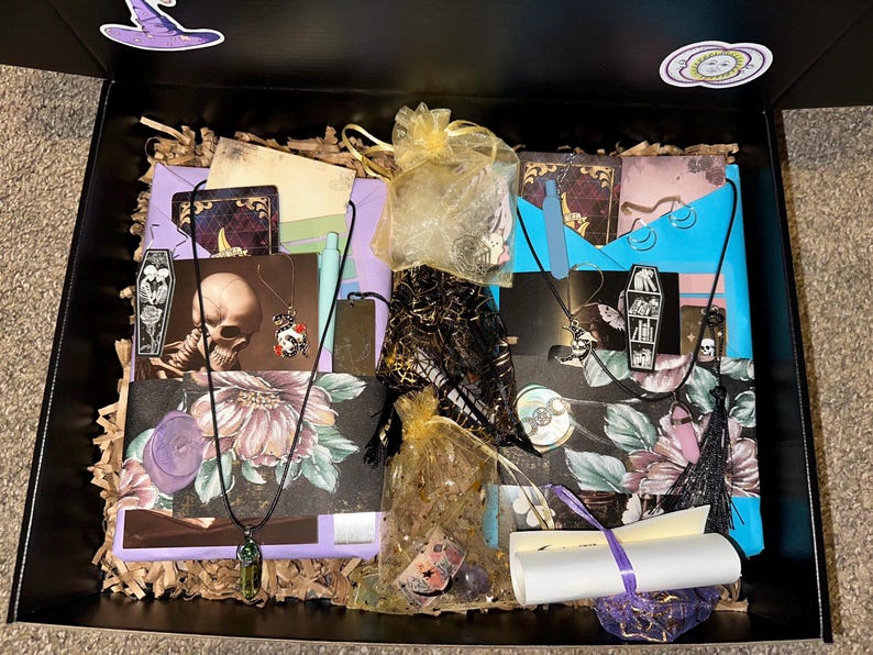 K&ouml;nnte beinhalten: Eine schwarze Box gef&uuml;llt mit Gothic-Artikeln. Die Box enth&auml;lt Halsketten, Karten mit Sch&auml;del- und Blumendesigns, kleine Beutel mit Kristallen und eine gerollte Schriftrolle, die mit einem lila Band gebunden ist. Dekorative Aufkleber eines Hexenhuts und einer Sonne befinden sich auf der Box.