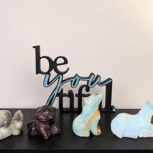 May include: A black sign with the text "be you tiful" in a light blue script font, positioned behind four carved stone animal figurines: a grey dog, a dark brown frog, a light blue wolf, and a translucent white horse.