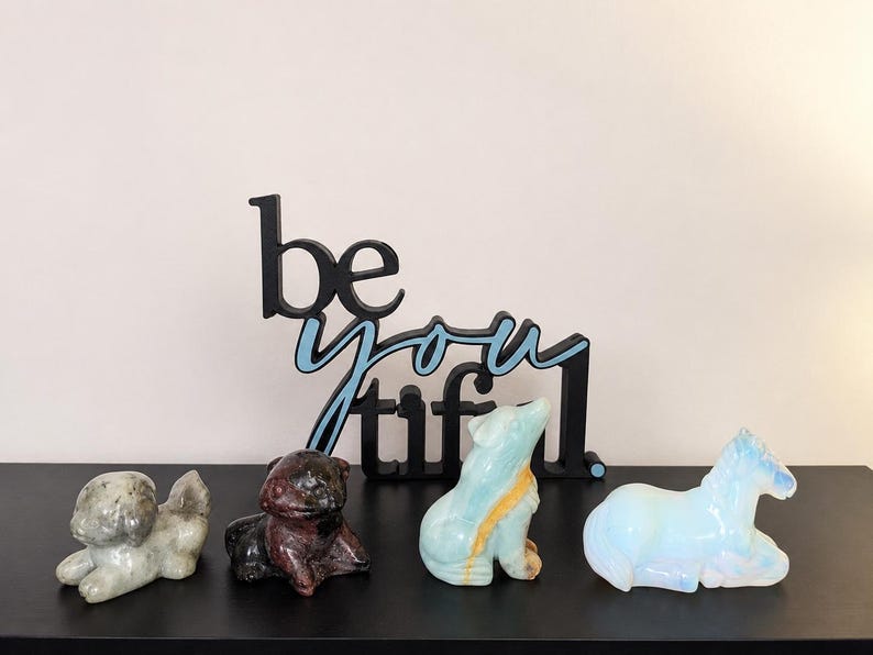 May include: A black sign with the text "be you tiful" in a light blue script font, positioned behind four carved stone animal figurines: a grey dog, a dark brown frog, a light blue wolf, and a translucent white horse.