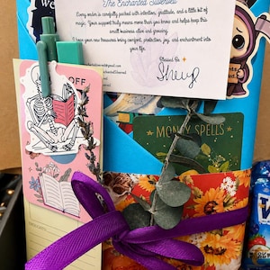 May include: A blue gift bag filled with assorted items including a thank you note from The Enchanted Silverveil, a skeleton sticker, a green pen, a sprig of eucalyptus, a purple ribbon, and a card labeled Money Spells with sunflower patterns.