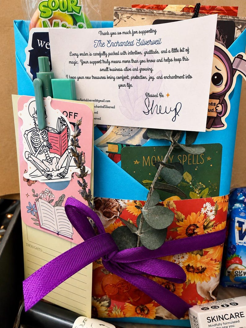 May include: A blue gift bag filled with assorted items including a thank you note from The Enchanted Silverveil, a skeleton sticker, a green pen, a sprig of eucalyptus, a purple ribbon, and a card labeled Money Spells with sunflower patterns.
