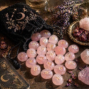 May include: A collection of pink crystal rune stones with gold symbols spilling from a black velvet bag with a crescent moon design. The stones are scattered on a wooden surface, with dried flowers and a small book visible.