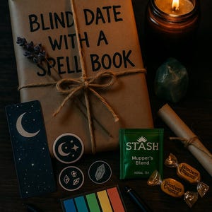 K&ouml;nnte beinhalten: Eine Anordnung auf dunklem Untergrund mit einem verpackten Buch mit der Aufschrift "BLIND DATE WITH A SPELL BOOK", einer brennenden Kerze, Aufklebern, Tee und S&uuml;&szlig;igkeiten. Die Szene erzeugt eine mystische Atmosph&auml;re.