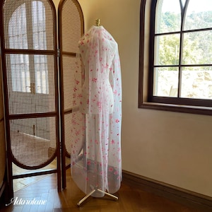 May include: A delicate white veil, adorned with pink floral embroidery, is displayed on a mannequin. The veil cascades down, showcasing the floral design. The mannequin is supported by a gold stand. The background features a wooden screen and a window.