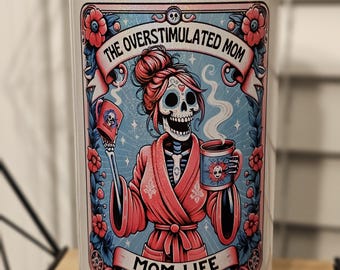May include: White ceramic tumbler with a glittery finish. The tumbler features a colorful illustration of a skeleton wearing a pink robe and holding a coffee mug. The text "The Overstimulated Mom" and "Mom Life" are printed on the tumbler.
